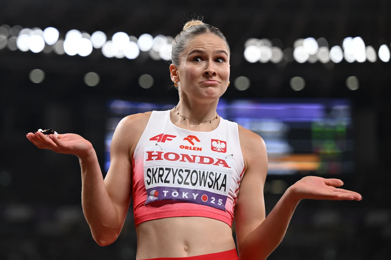 Pia Skrzyszowska Lekkoatletka w stroju sportowym podnosi ramiona w geście wyrażającym zaskoczenie lub bezradność, widoczna nazwa na stroju i tło stadionu lekkoatletycznego.