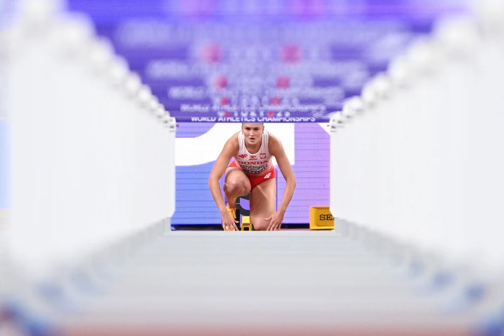 Pia Skrzyszowska Lekkoatletka w stroju startowym pochylona w pozycji startowej na bieżni stadionu, w tle tablica z nazwą zawodów i jaskrawe światło, po bokach rozmyte elementy konstrukcji stadionowej tworzą tunelowy efekt kadru.