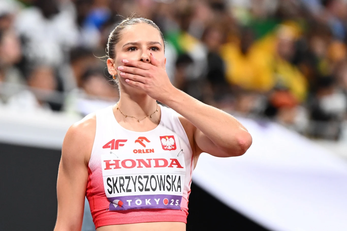 Lekkoatletka w stroju sportowym, z ręką przy ustach, na tle rozmytej publiczności, widoczny numer startowy i nazwisko Skrzyszowska na koszulce.