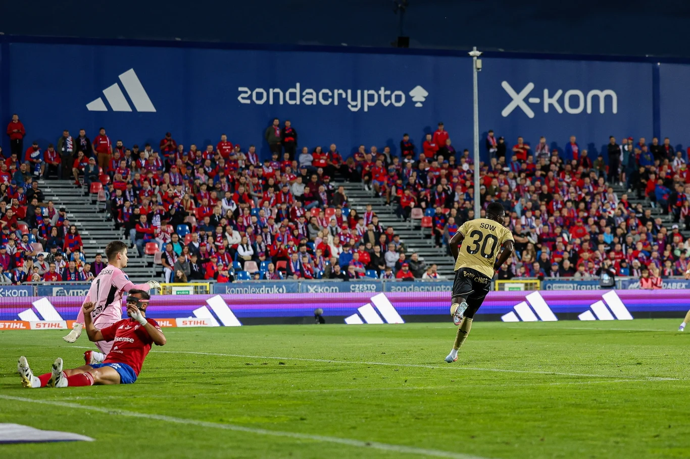 Ousmane Sow świętujący gola dla Górnika Zabrze w starciu z Rakowem Częstochowa Piłkarze w trakcie meczu walczą o piłkę na murawie stadionu, w tle trybuny pełne kibiców ubrane głównie na czerwono, a za nimi widoczne są duże reklamy firm partnerskich.
