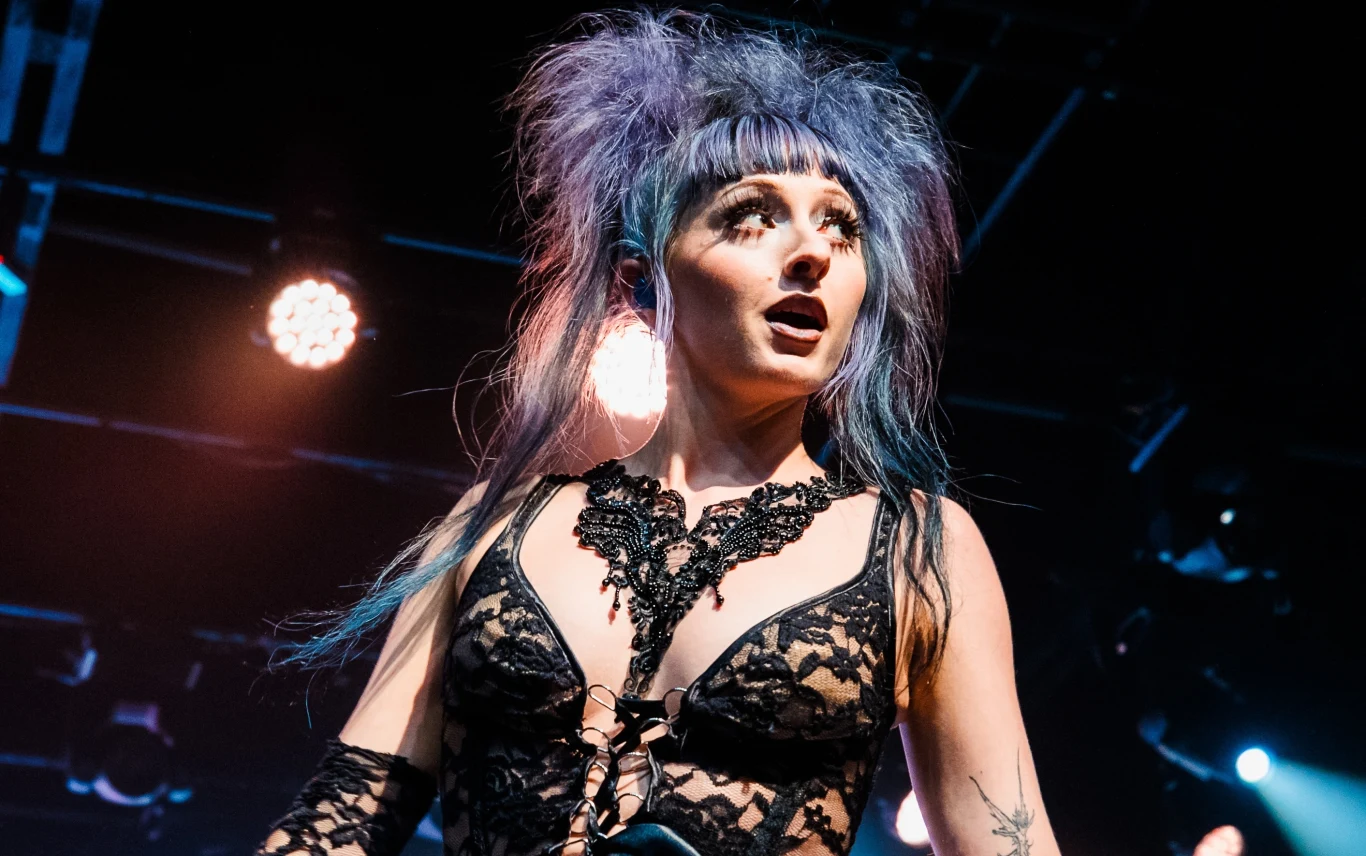 Ashnikko powraca do Polski Kobieta z wyrazistym makijażem i fioletowymi włosami, ubrana w czarny koronkowy strój sceniczny, stoi na jasno oświetlonej scenie, tło stanowią reflektory i ciemność sali koncertowej