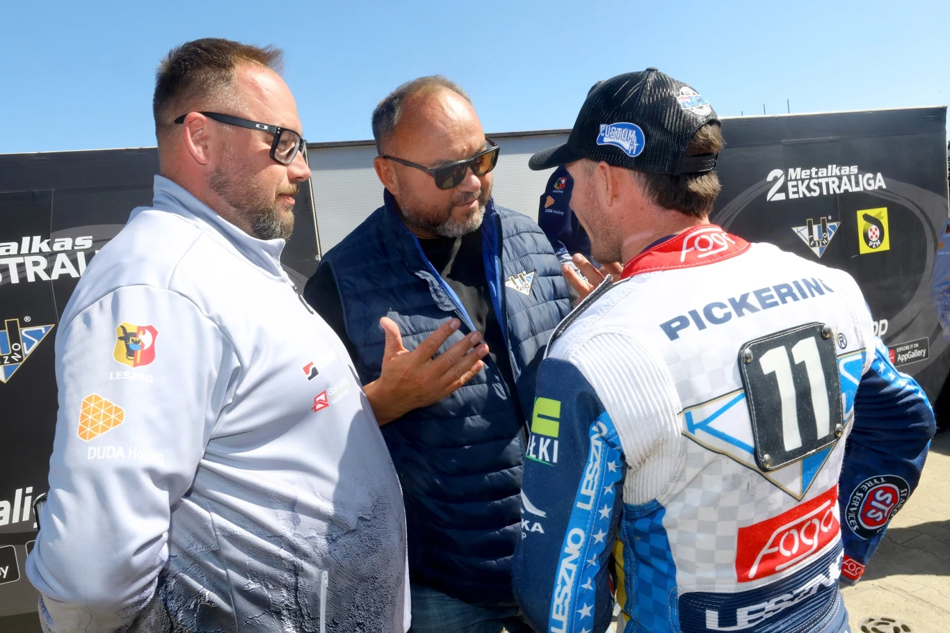 Trzech mężczyzn w rozmowie podczas wydarzenia sportowego, jeden z nich ubrany w kombinezon żużlowy z numerem 11 i napisem 'Pickering', pozostali mężczyźni w sportowych kurtkach, w tle banery sponsorów ligi żużlowej.