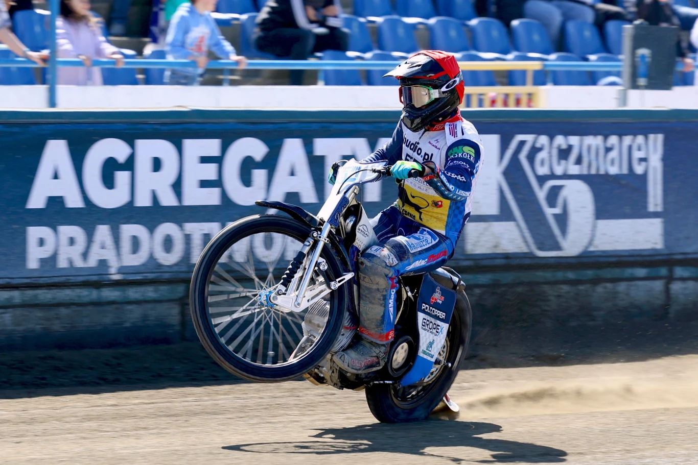 Zawodnik żużlowy w kolorowym kombinezonie wykonuje efektowny wheelie podczas wyścigu na torze, wokół puste niebieskie trybuny i widoczne napisy sponsorów na bandzie.