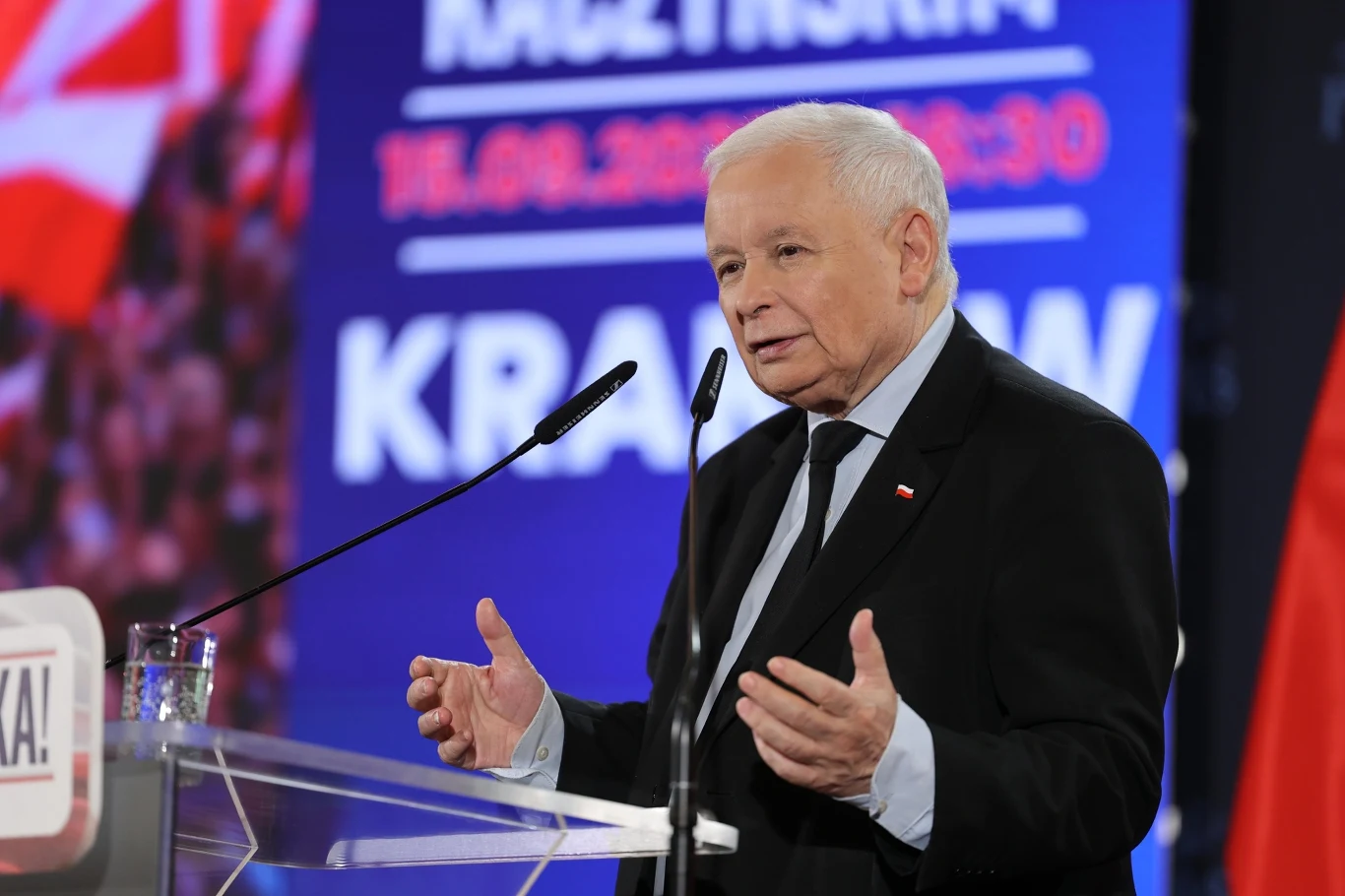 Prezes PiS Jarosław Kaczyński na spotkaniu z sympatykami Starszy mężczyzna stojący przy mównicy, wygłaszający przemówienie, w tle widoczne flagi oraz niebieski ekran z napisem i datą.