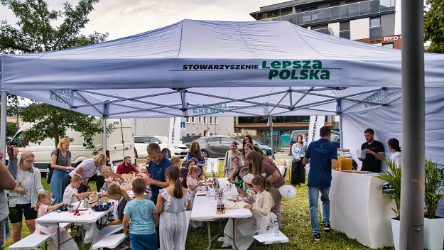 Ludzie, dorośli i dzieci, zgromadzeni pod dużym białym namiotem organizacji Lepsza Polska, uczestniczą w warsztatach plastycznych i spotkaniach, wokół zielona trawa i nowoczesne zabudowania w tle.