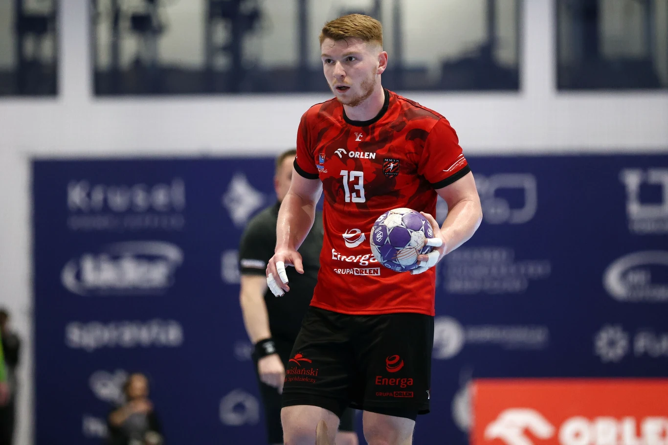 Zawodnik piłki ręcznej w czerwonej koszulce z numerem trzynaście, trzymający piłkę w rękach, skoncentrowany podczas meczu, na tle niebieskich band reklamowych i hali sportowej.