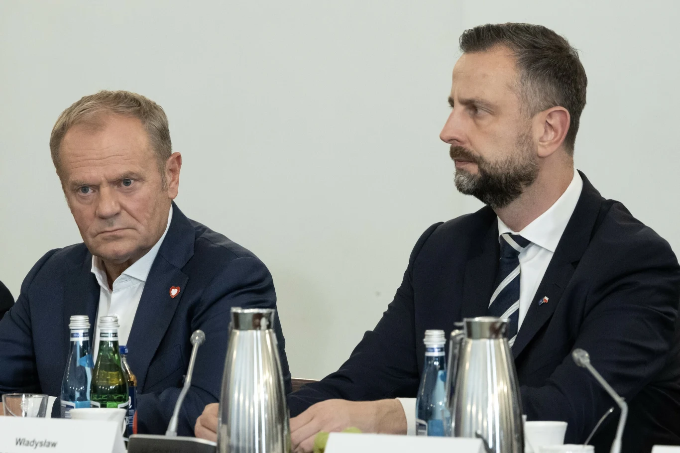 Donald Tusk i Władysław Kosiniak-Kamysz Dwaj mężczyźni w garniturach siedzą obok siebie przy stole konferencyjnym, otoczeni butelkami wody, karafkami oraz tabliczkami z imionami. Obaj wyglądają na skupionych i zaangażowanych w rozmowę.