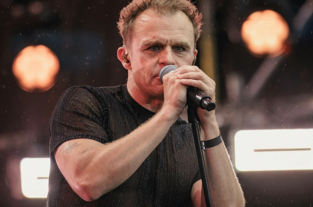 Piotr Rogucki współtworzy projekt "Światło i MRock" z Katarzyną Nosowską Mężczyzna śpiewający do mikrofonu na scenie podczas występu, tło rozmyte z widocznymi światłami sceny, silna ekspresja oraz skupienie na twarzy artysty.