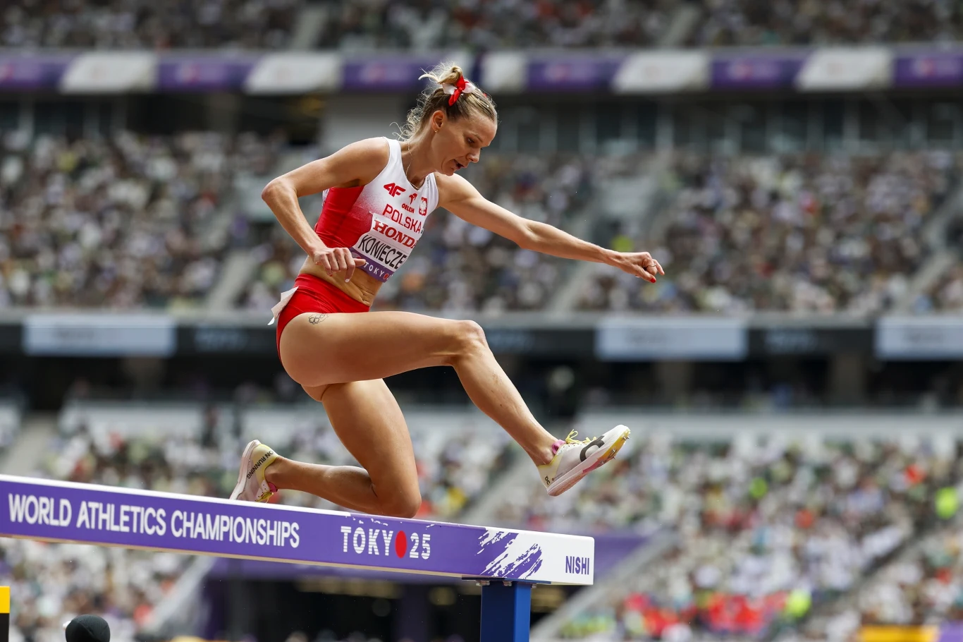 Alicja Konieczek Zawodniczka w biało-czerwonym stroju startowym przeskakuje przeszkodę podczas biegu przez płotki na stadionie lekkoatletycznym, w tle widoczna publiczność oraz tablice z nazwą zawodów Mistrzostw Świata w lekkoatletyce w Tokio.