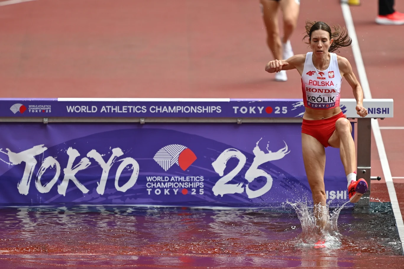 Kinga Królik Zawodniczka reprezentująca Polskę podczas biegu z przeszkodami na Mistrzostwach Świata w Lekkoatletyce w Tokio, przeskakująca przez wodny rów na stadionie lekkoatletycznym.
