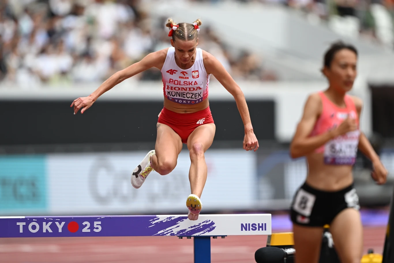 Alicja Konieczek Zawodniczka reprezentacji Polski pokonuje przeszkodę podczas biegu na stadionie lekkoatletycznym, obok biegnie rywalka w różowej koszulce, w tle rozmazana publiczność.