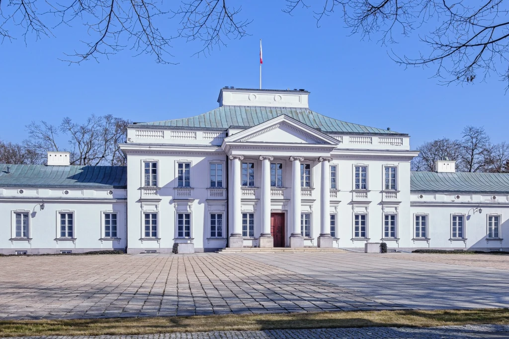 Neoklasycystyczny pałac o jasnej elewacji z kolumnowym portykiem, wysoka mansardowa część centralna, po bokach dwa niższe skrzydła, przed budynkiem szeroki brukowany plac, na dachu powiewa flaga państwowa.
