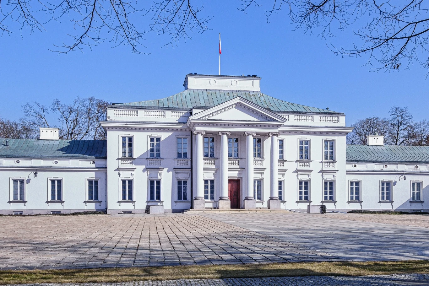 Belweder Neoklasycystyczny pałac o jasnej elewacji z kolumnowym portykiem, wysoka mansardowa część centralna, po bokach dwa niższe skrzydła, przed budynkiem szeroki brukowany plac, na dachu powiewa flaga państwowa.