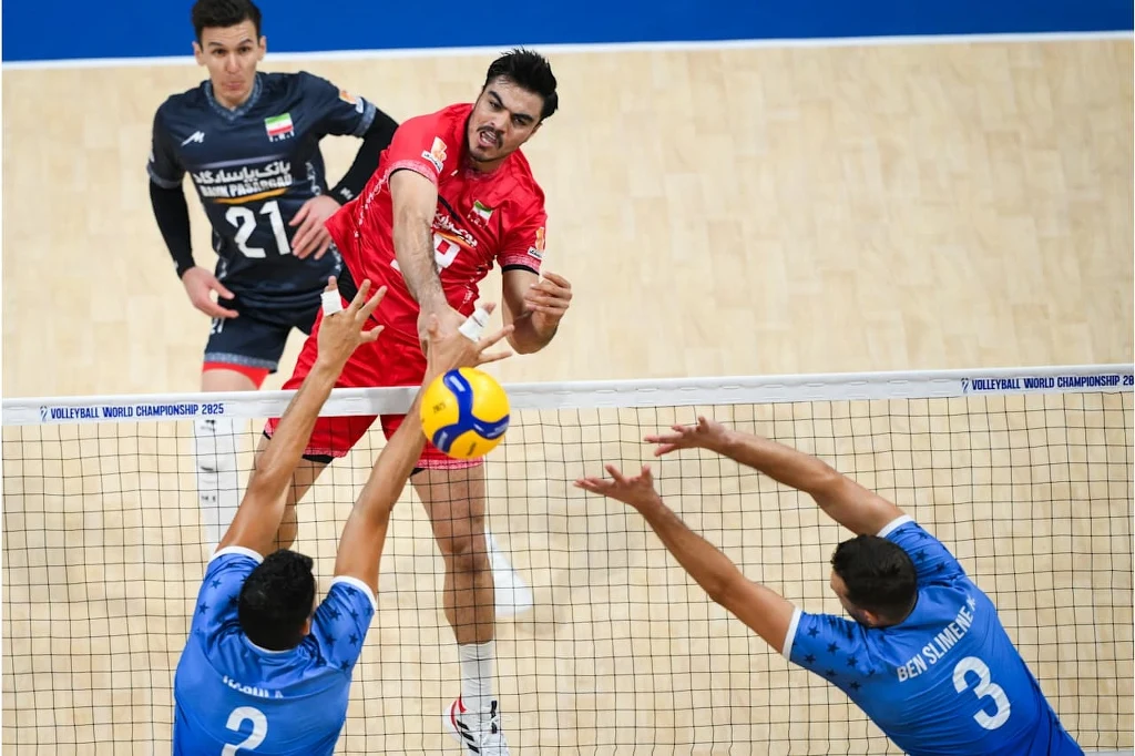 MŚ siatkarzy. Sharifi Morteza atakuje w meczu Iran - Tunezja Siatkarz w czerwonym stroju wykonuje atak ponad siatką, próbując przebić piłkę przez blok dwóch przeciwników ubranych w niebieskie stroje, w tle widoczny zawodnik drużyny w ciemnym stroju.