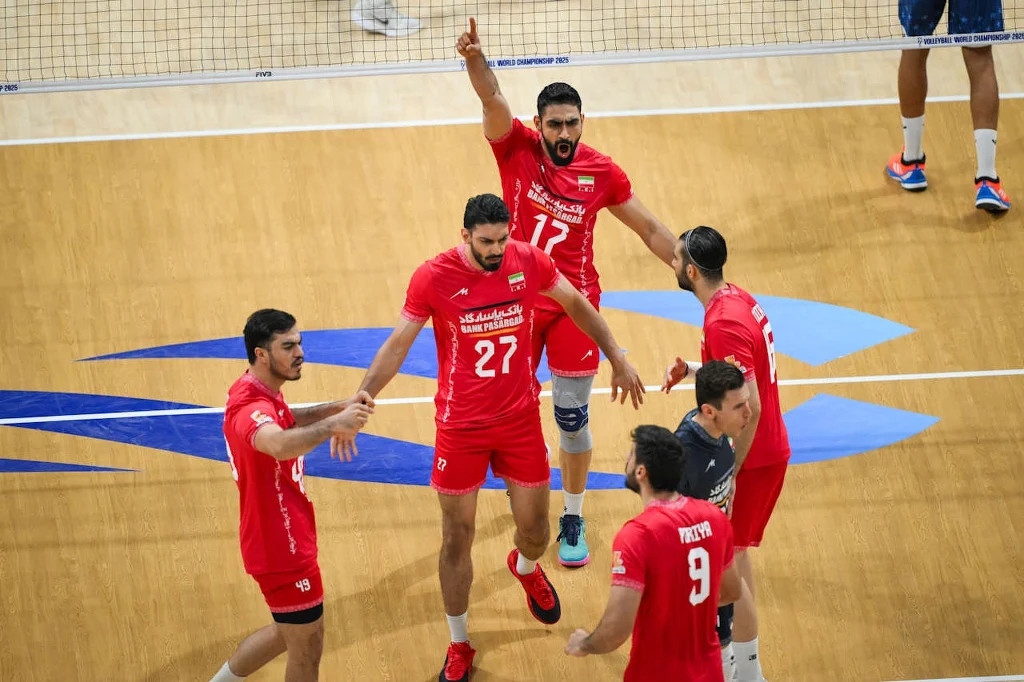 Siatkarze reprezentacji Iranu Grupa siatkarzy w czerwonych strojach świętuje zdobycie punktu na boisku do siatkówki, kilku zawodników unosi ręce w geście radości, w tle widoczna siatka, dominują kolory niebieski oraz żółty parkietu sportowego.