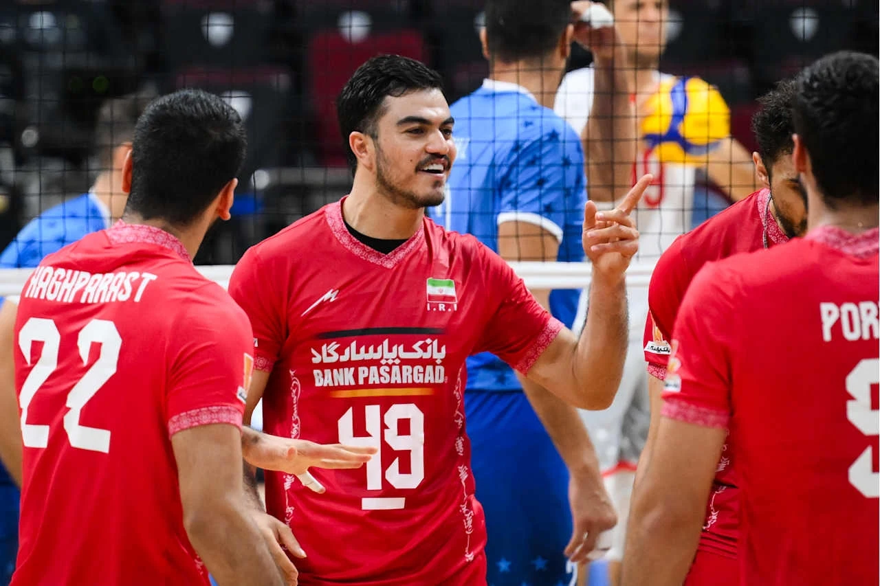 Grupa siatkarzy w czerwonych strojach drużyny narodowej świętuje punkt podczas meczu, na pierwszym planie zawodnik z numerem 49 gestykuluje energetycznie, wokół niego widać innych członków drużyny i fragment siatki.
