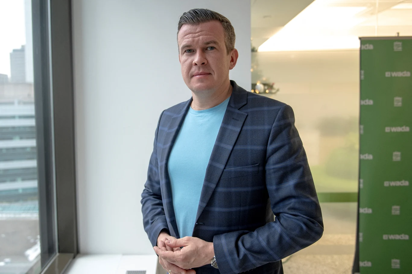 Mężczyzna w marynarce w niebieską kratę i jasnoniebieskiej koszulce stoi obok okna, w tle rozmazana panorama miasta oraz zielony baner z logo.