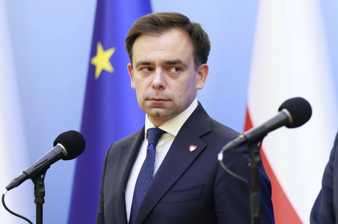 Domański o KSEF. "Działa płynnie, coraz więcej osób się loguje" Mężczyzna w garniturze i krawacie stoi na tle flag Unii Europejskiej i Polski, skierowany lekko w bok, z poważnym wyrazem twarzy, przed mikrofonami podczas konferencji lub oficjalnego wystąpienia.