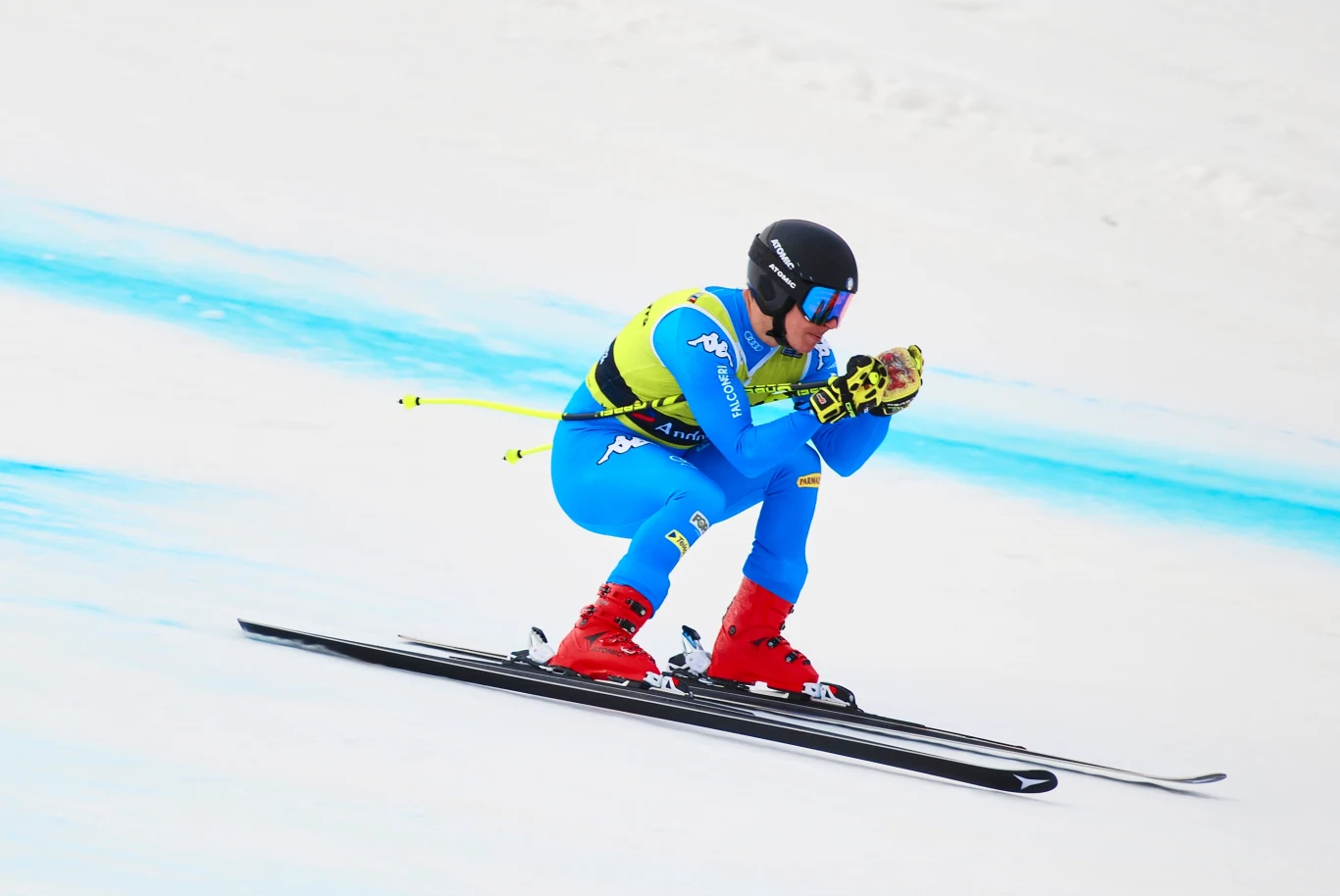 Narciarz w niebieskim kombinezonie sportowym i kasku podczas zjazdu na nartach po zaśnieżonym stoku, dynamiczna pozycja zjazdowa, wyraziste czerwone buty narciarskie.