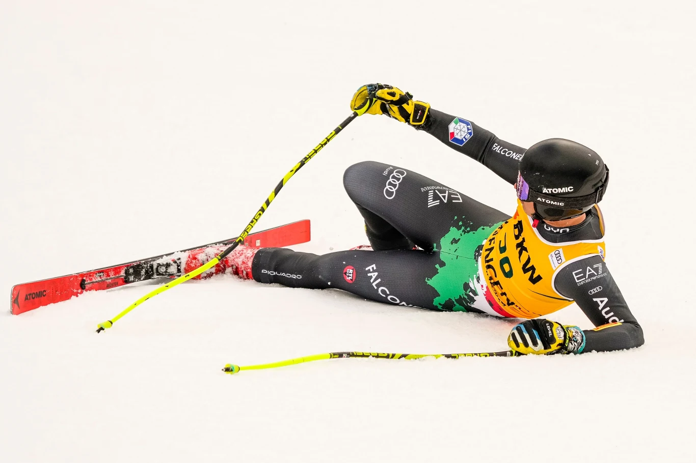 Narciarz alpejski w sportowym kombinezonie leży na śniegu po upadku podczas zawodów, trzymając kijki narciarskie w rękach, wokół biały śnieg, widać wyraźnie narty oraz numer startowy na piersi.