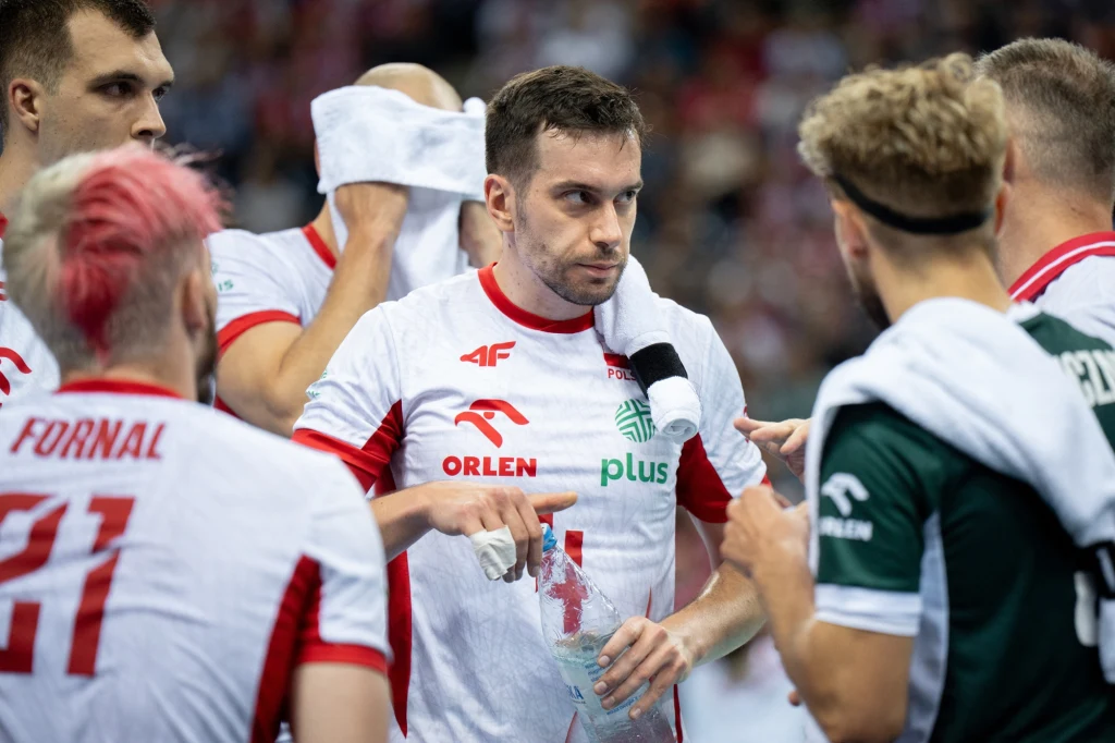 Marcin Komenda (w środku kadru) Zawodnicy drużyny siatkarskiej w trakcie narady na boisku, skupieni wokół jednego z graczy, w tle rozmyta publiczność.