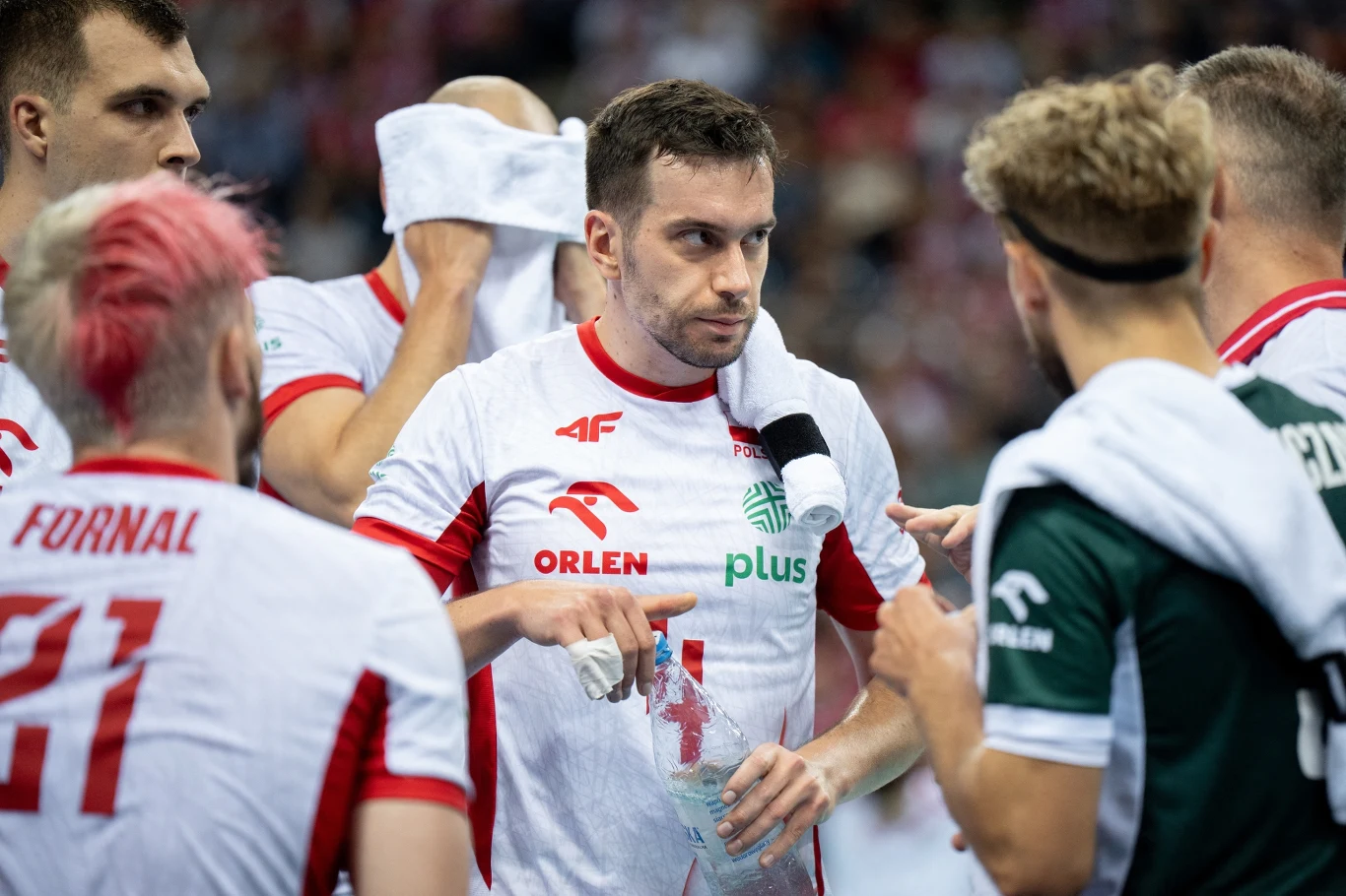 Zawodnicy drużyny siatkarskiej w trakcie narady na boisku, skupieni wokół jednego z graczy, w tle rozmyta publiczność.