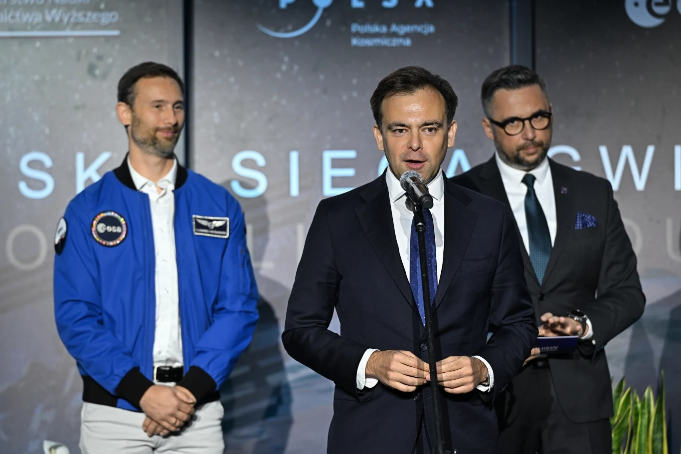 Sławosz Uznański-Wiśniewski rusza w trasę naukowo-technologiczną po Polsce Trzech mężczyzn w oficjalnych strojach stoi na tle planszy z logotypami instytucji, jeden z nich przemawia do mikrofonu, obok niego mężczyzna w niebieskiej kurtce z naszywką ESA oraz mężczyzna w garniturze z notatnikiem.