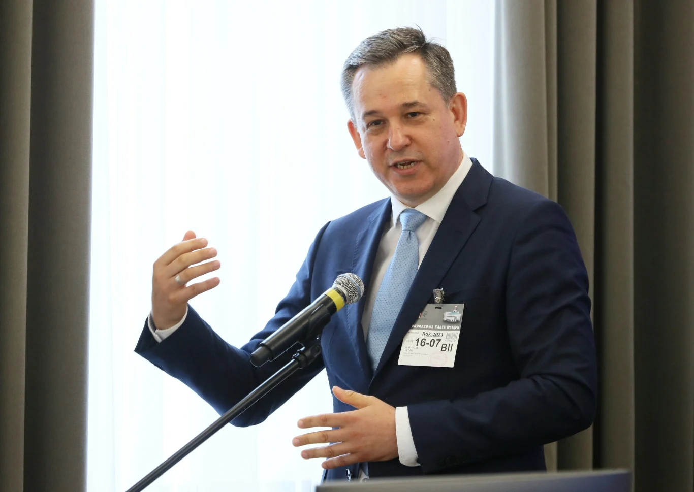 Mężczyzna w garniturze przemawia przy mikrofonie w sali konferencyjnej, na marynarce ma przypiętą identyfikacyjną plakietkę i gestykuluje ręką podkreślając wypowiedź, w tle widoczne zasłony i światło wpadające przez okno.