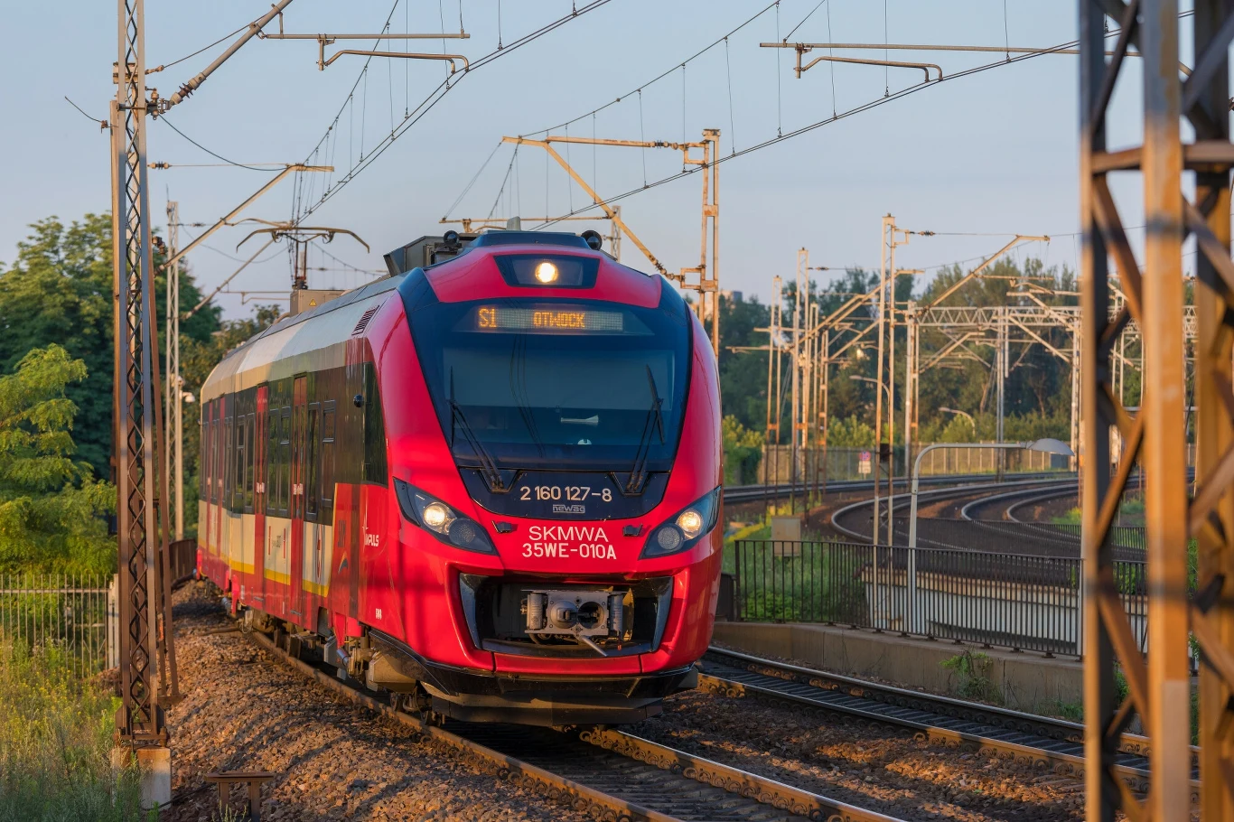 Poważne utrudnienia na warszawskich torach. ZTM z komunikatem Czerwony elektryczny pociąg pasażerski poruszający się po zakrzywionych torach kolejowych, otoczony infrastrukturą trakcyjną i zielenią, w naturalnym świetle dziennym.