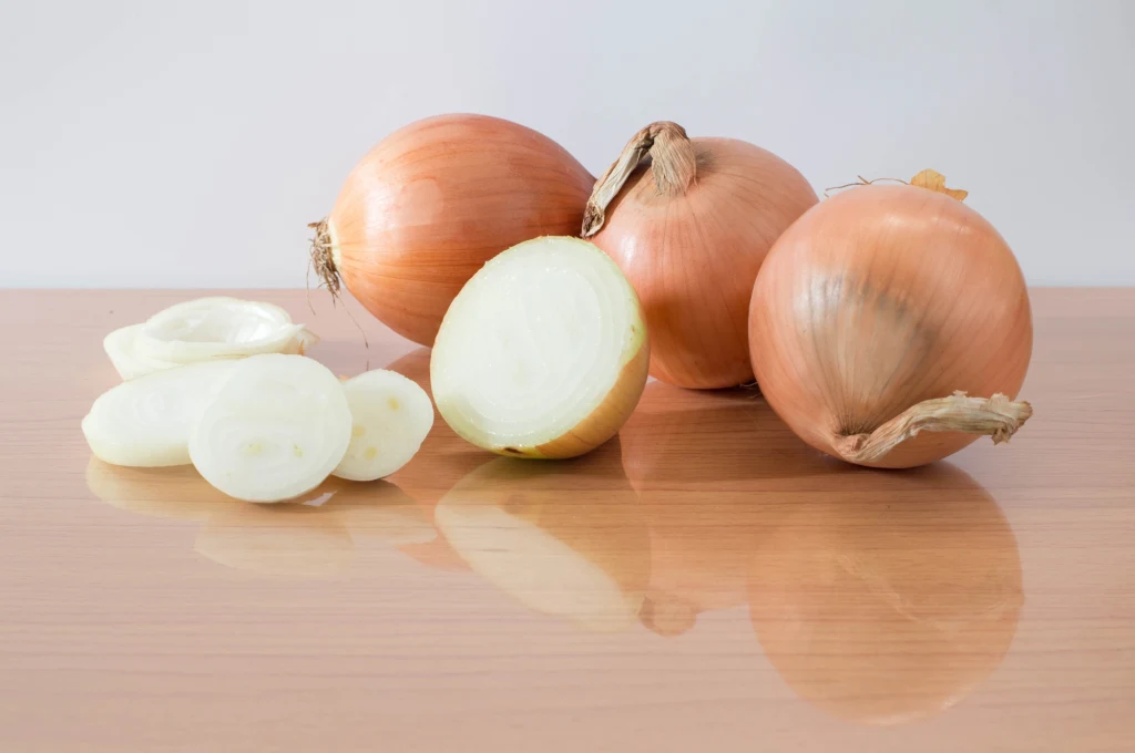 Trzy żółte cebule oraz jedna pokrojona cebula leżące na gładkiej, drewnianej powierzchni, jasne tło.