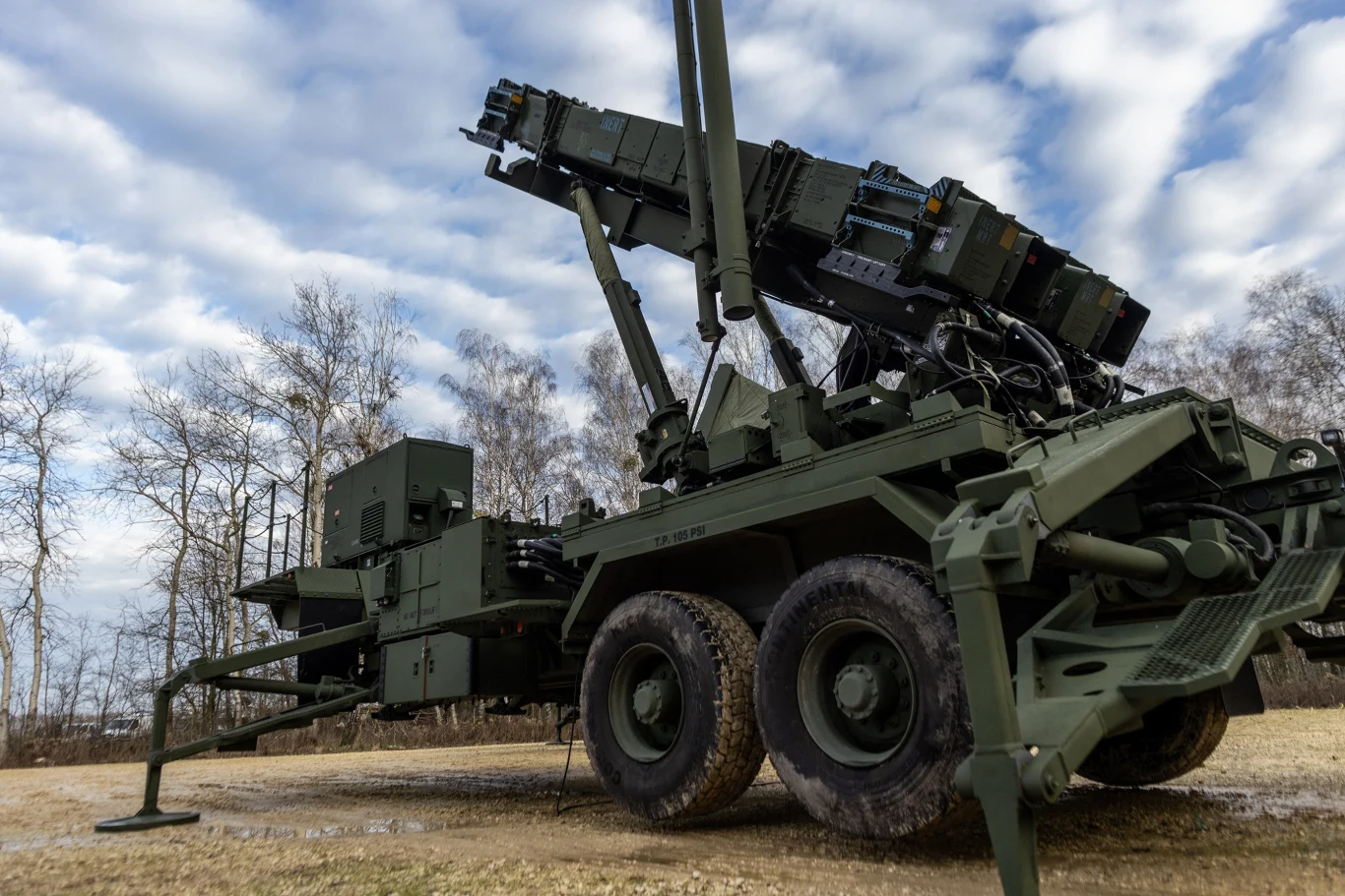 Wojskowy zestaw rakietowy ustawiony na podwoziu ciężarówki na otwartym terenie, otoczony drzewami bez liści oraz niebem pokrytym chmurami.