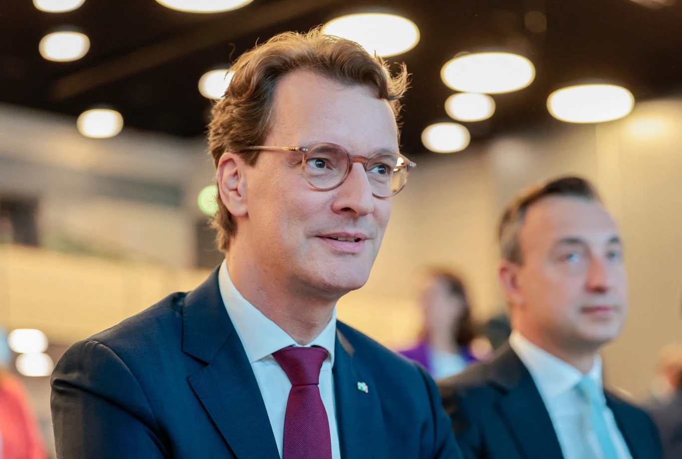 Szef niemiecko-polskiej grupy parlamentarnej w Bundestagu Paul Ziemiak Mężczyzna w garniturze i okularach siedzi w dobrze oświetlonym pomieszczeniu, w tle drugi mężczyzna w garniturze, rozmyte postacie oraz jasne, okrągłe lampy sufitowe.