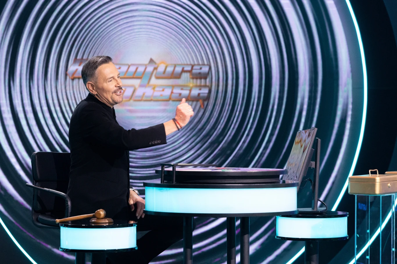 "Awantura o kasę". Teleturniej w Polsacie o 17:30 w sobotę i online na Polsat Box Go Mężczyzna w średnim wieku siedzi przy nowoczesnym, okrągłym biurku w studiu telewizyjnym, na tle dużego ekranu z napisem Znajdź Wspólną Pasję, gestykuluje z entuzjazmem i uśmiecha się, obok widoczny jest młotek sędziowski.