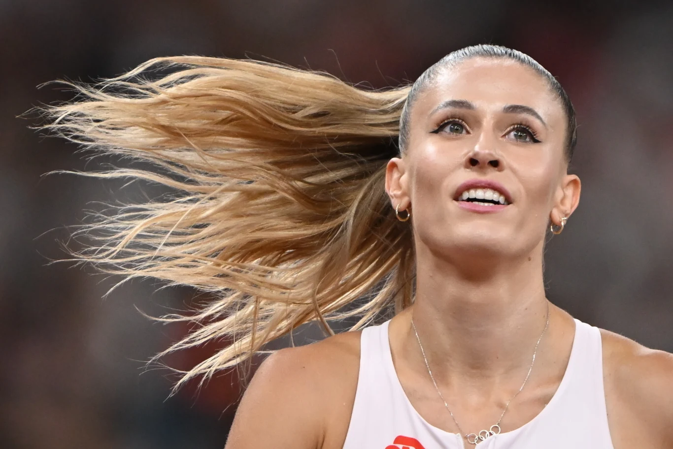 Lekkoatletka w białym stroju sportowym na stadionie, z widocznym ruchem rozwianych blond włosów i wyrazem skupienia na twarzy.