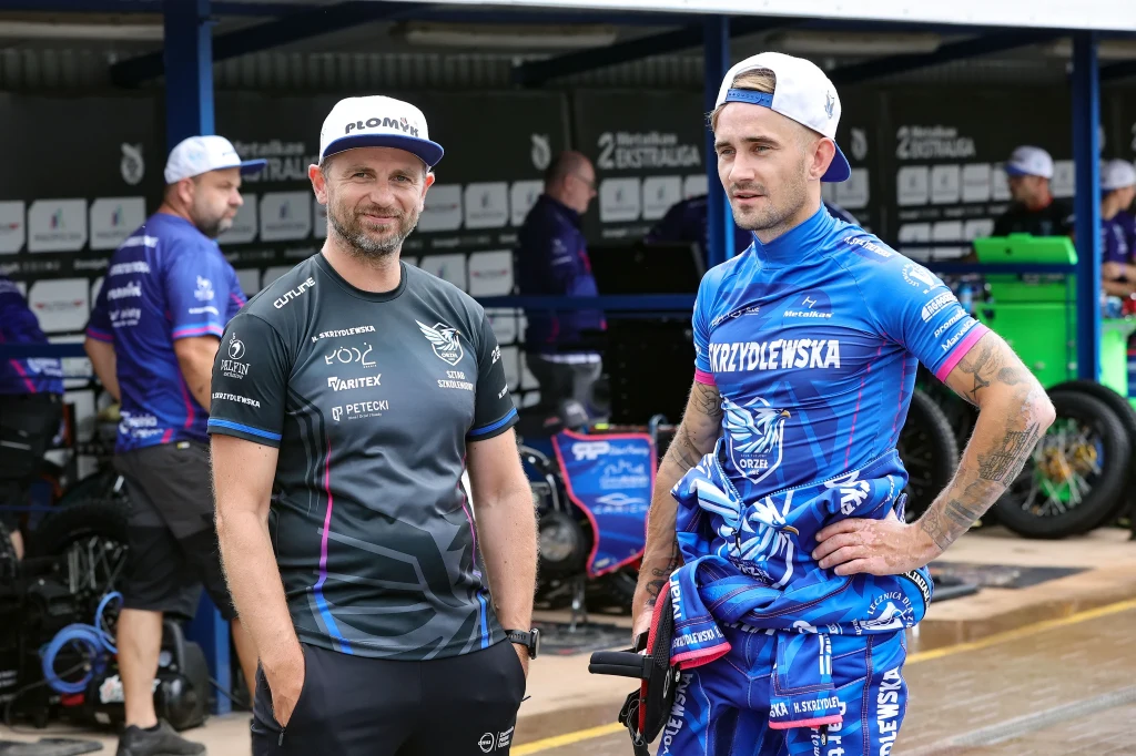 Dwóch mężczyzn ubranych w stroje sportowe do żużlu stoi obok siebie w strefie serwisowej toru żużlowego; w tle ludzie przy motocyklach oraz bannery z nazwami sponsorów.