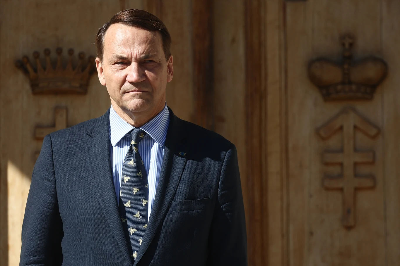 Radosław Sikorski odpowiedział na zaczepkę prawicowego publicysty Mężczyzna w garniturze i pod krawatem stoi na tle drewnianych drzwi ozdobionych koronami i symbolami