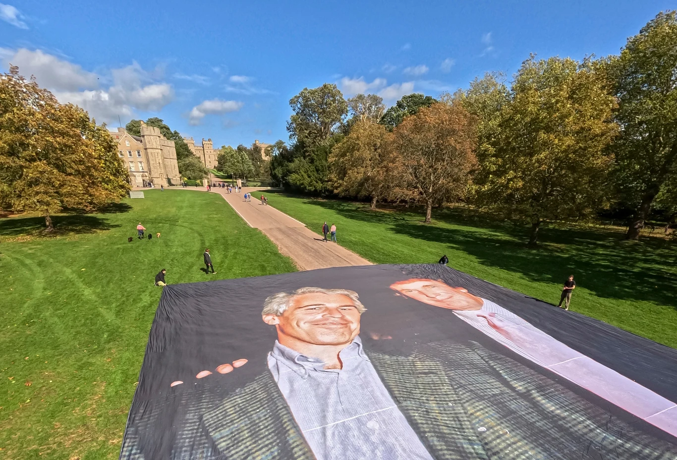 Przed Zamkiem Windsor rozciągnięto zdjęcie Donalda Trumpa i Jeffreya Epsteina Ogromny baner z wydrukowanymi wizerunkami dwóch mężczyzn rozłożony na trawniku w dużym parku, w tle widoczna aleja prowadząca w kierunku zamku otoczonego drzewami.