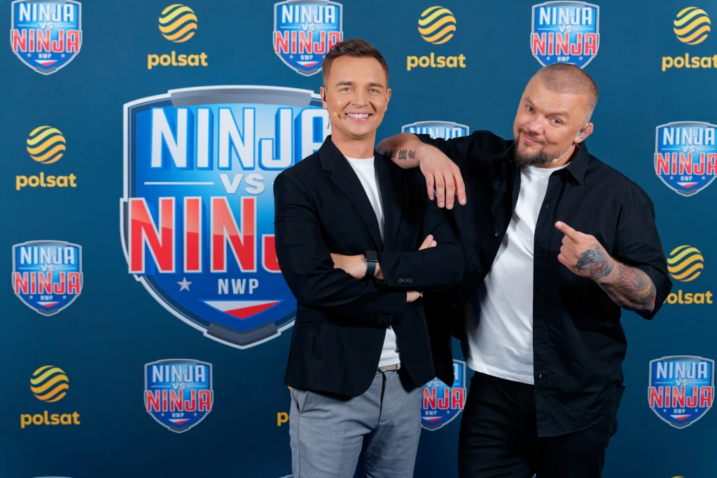 Przed nami trzeci odcinek „Ninja vs Ninja” Dwóch mężczyzn pozuje na tle ścianki promującej program Ninja vs Ninja z widocznymi logotypami Polsatu, obaj uśmiechnięci w nieformalnych pozach.