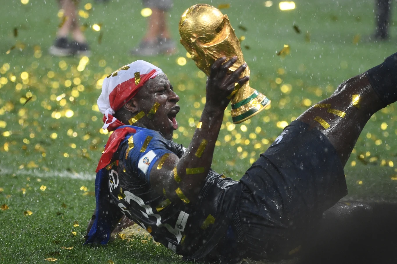 Benjamin Mendy Piłkarz w stroju reprezentacji Francji leży na murawie i triumfalnie unosi trofeum mistrzostw świata, wokół niego pada deszcz złotego konfetti, a na jego głowie widnieje opaska w narodowych barwach.