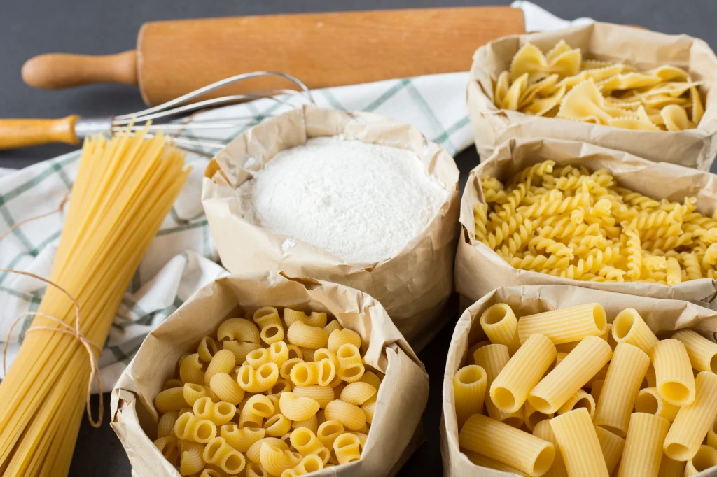 Różne rodzaje makaronów ułożone w papierowych torebkach obok mąki, trzepaczki i wałka do ciasta na tle kuchennej ściereczki.