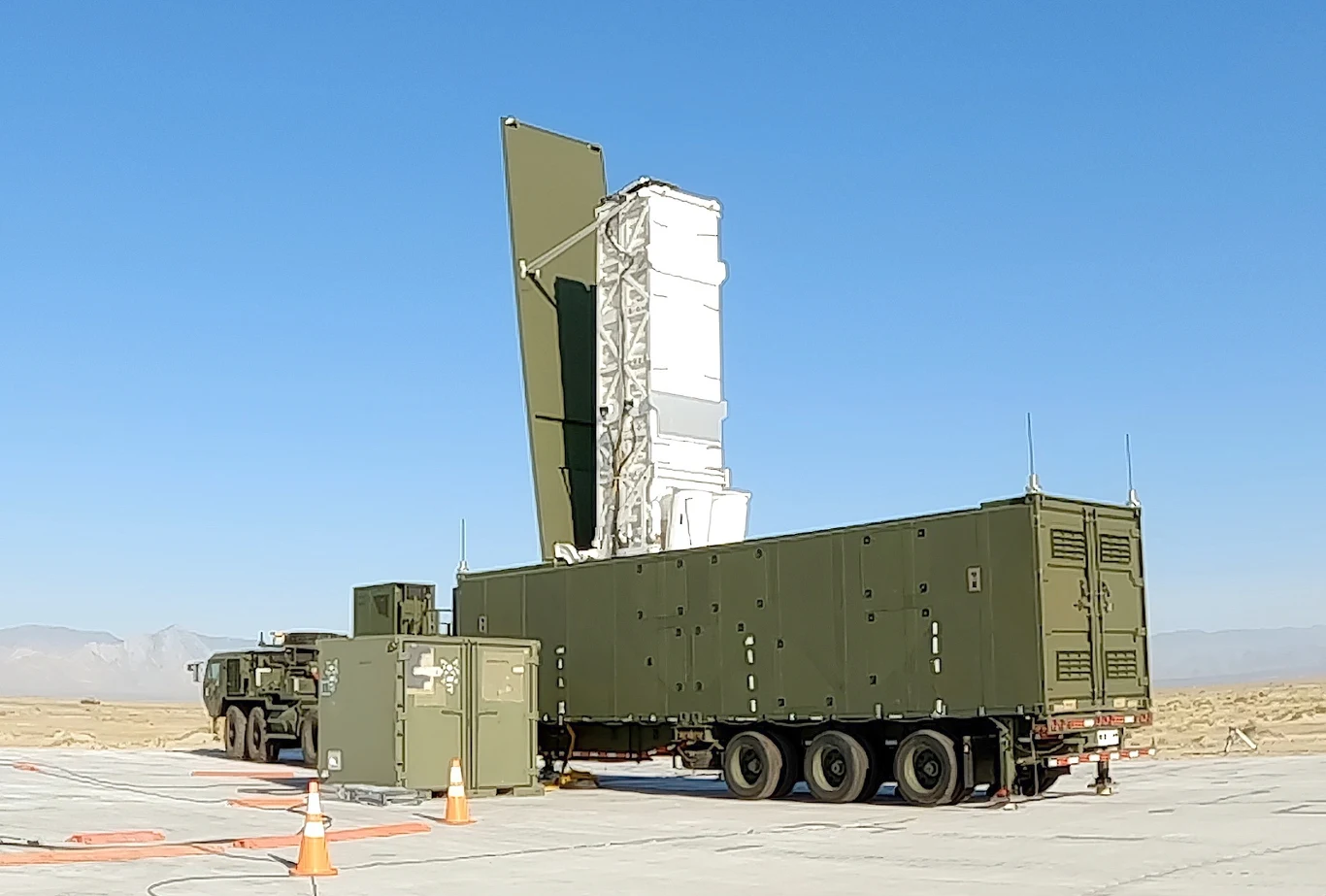 Wojskowy system radarowy lub komunikacyjny dużych rozmiarów ustawiony na utwardzonym placu z widocznymi elementami technicznymi, na tle pustynnego krajobrazu i jasnego nieba.