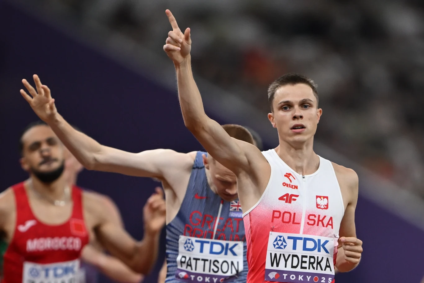 Lekkoatleta w stroju reprezentacji Polski wiwatuje po ukończonym biegu na hali, obok widoczni są zawodnicy z innych krajów, wokół dynamiczna atmosfera sportowej rywalizacji.