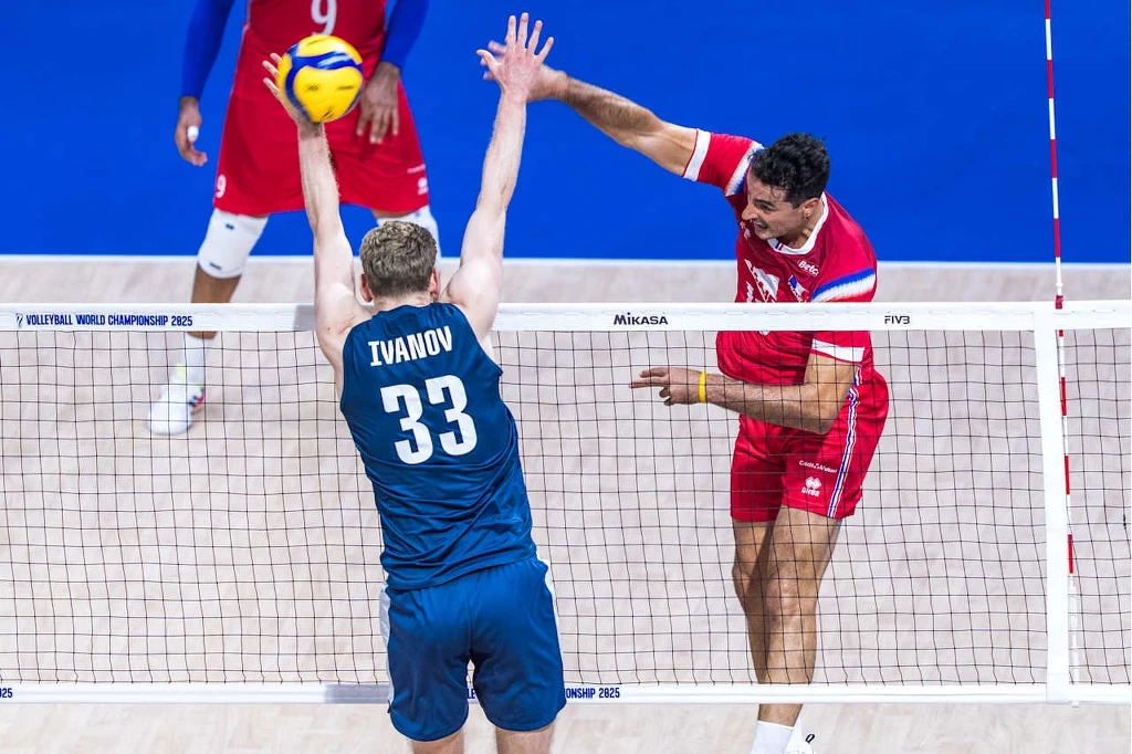 Siatkówka. Yacine Louati atakuje w meczu Francja - Finlandia Dwóch siatkarzy w akcji po obu stronach siatki podczas meczu siatkówki, zawodnik w niebieskim stroju z numerem 33 próbuje zablokować atak gracza w czerwonym stroju, nad siatką unosi się piłka.