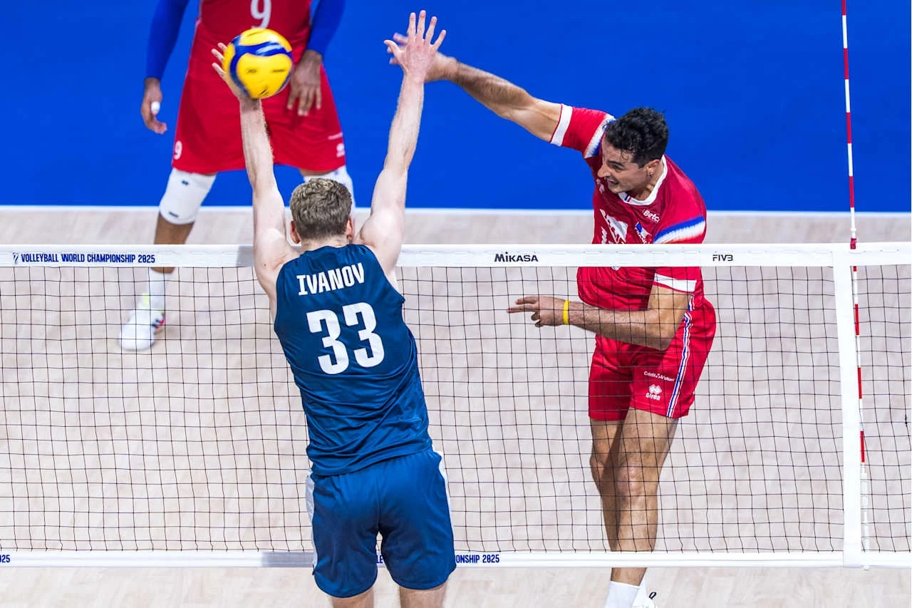 Dwóch siatkarzy w akcji po obu stronach siatki podczas meczu siatkówki, zawodnik w niebieskim stroju z numerem 33 próbuje zablokować atak gracza w czerwonym stroju, nad siatką unosi się piłka.