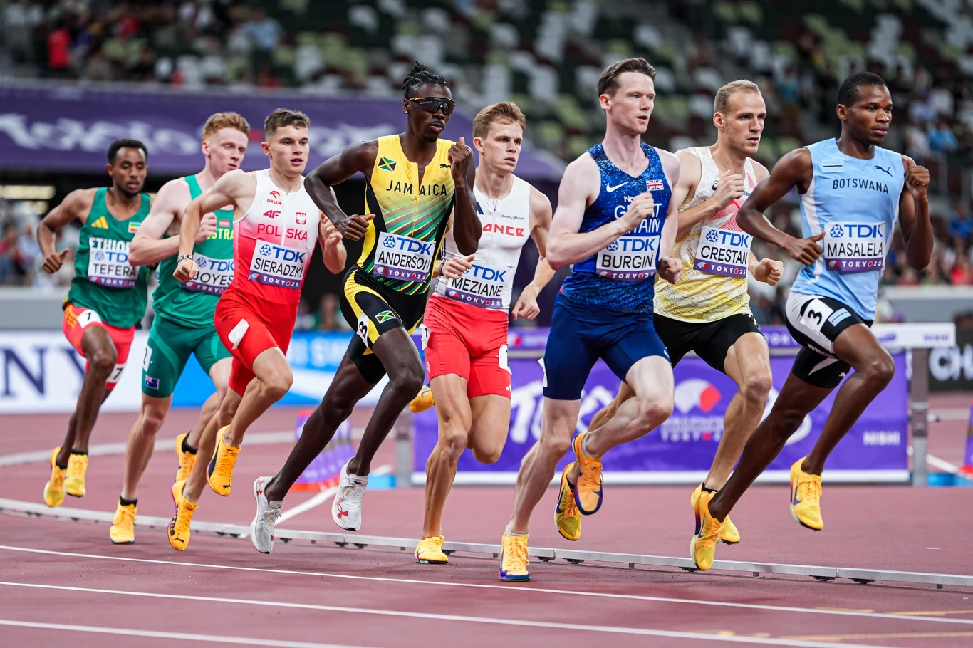 Patryk Sieradzki w Tokio Grupa zawodników biegnących po torze lekkoatletycznym podczas zawodów, ubranych w stroje sportowe reprezentujące różne kraje, widoczne intensywne emocje i rywalizacja.