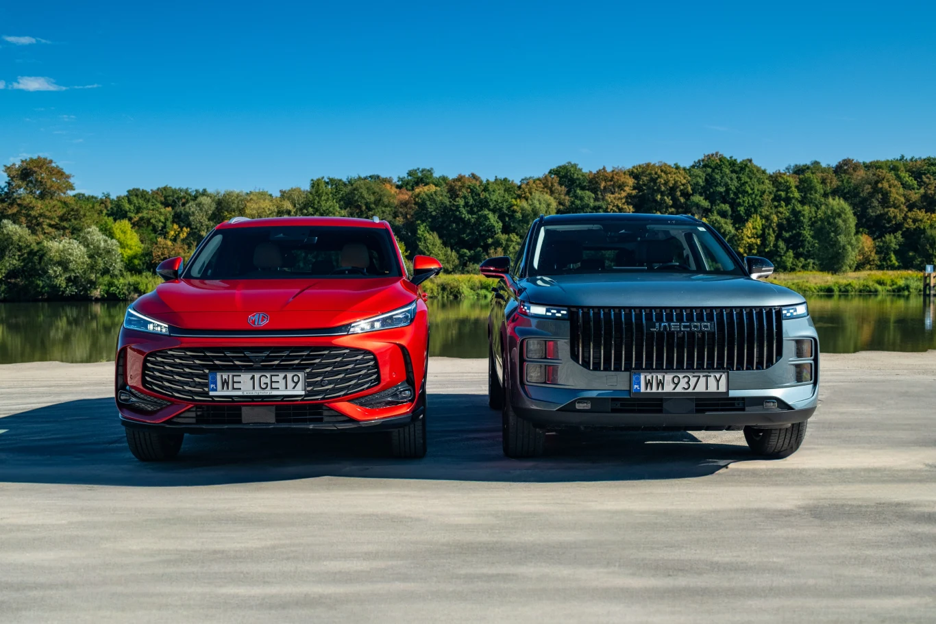 Dwa nowoczesne samochody SUV, czerwony i szary, ustawione obok siebie na tle zielonych drzew oraz jeziora, jasne niebo bez chmur.