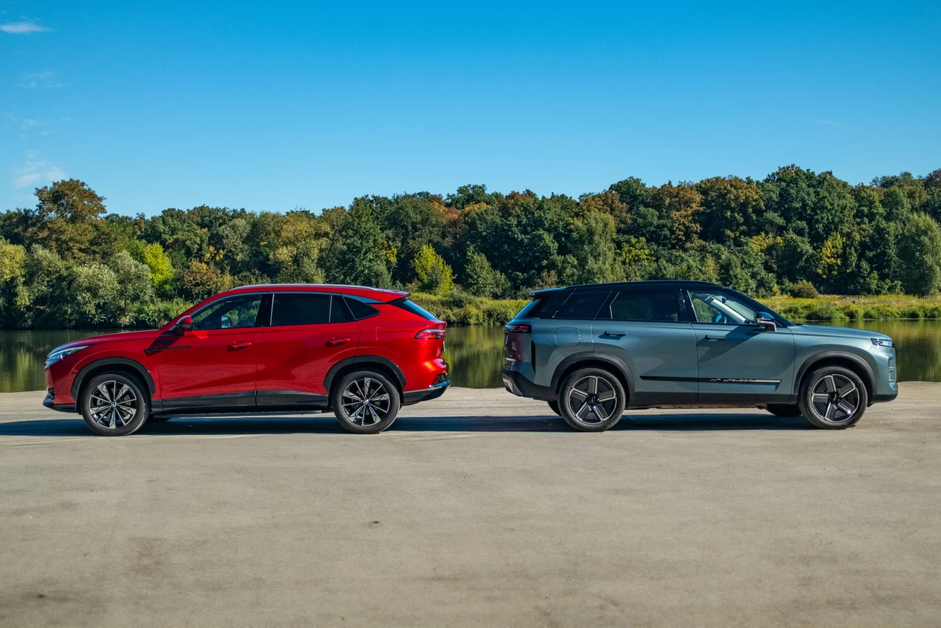 Dwa nowoczesne SUV-y stojące obok siebie na betonowej nawierzchni, w tle gęsty zielony las i niebieskie niebo bez chmur. Samochody różnią się kolorem, jeden jest czerwony, drugi niebiesko-szary.