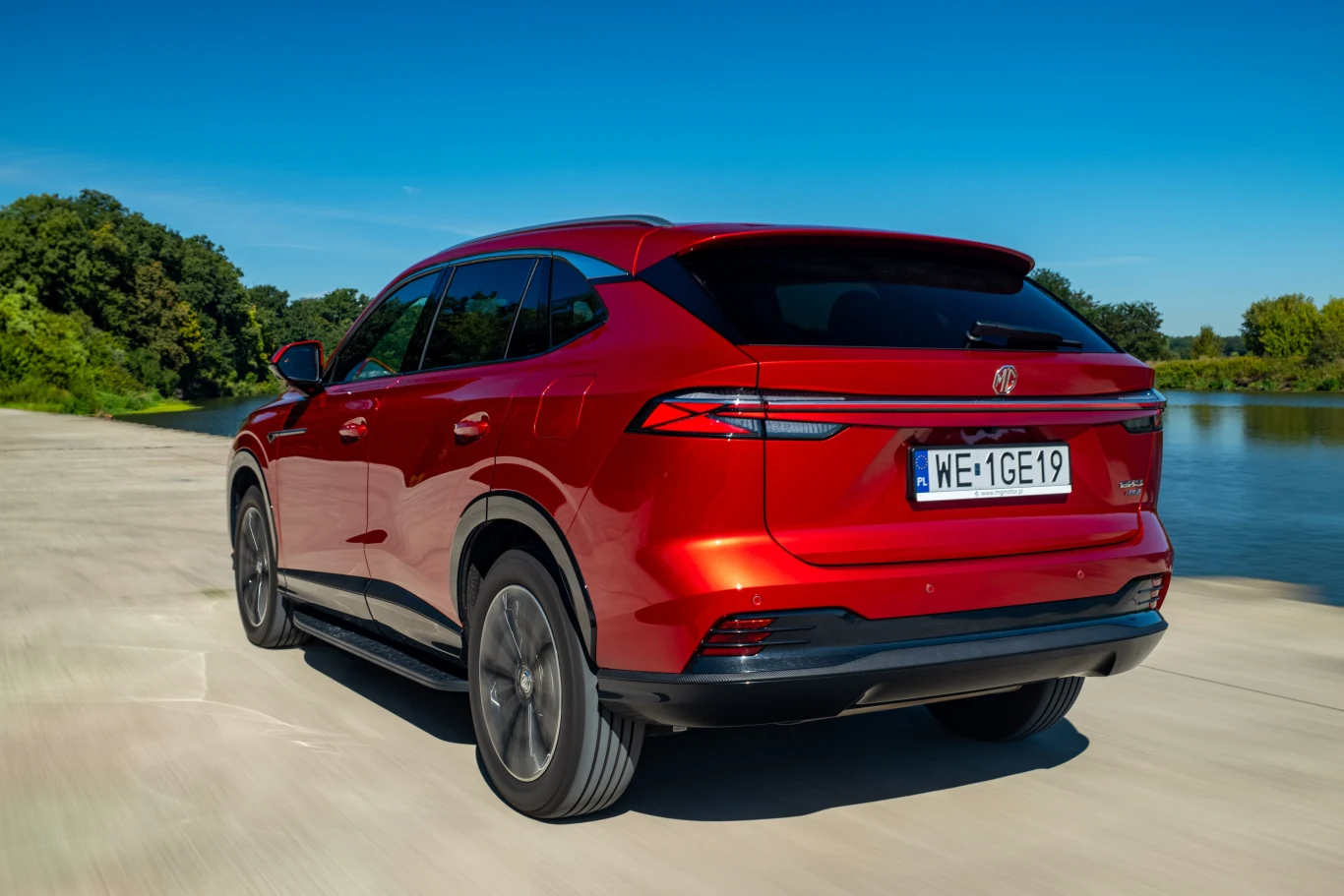 Czerwony nowoczesny SUV na asfaltowej drodze, w tle jezioro oraz zielone drzewa pod błękitnym niebem.