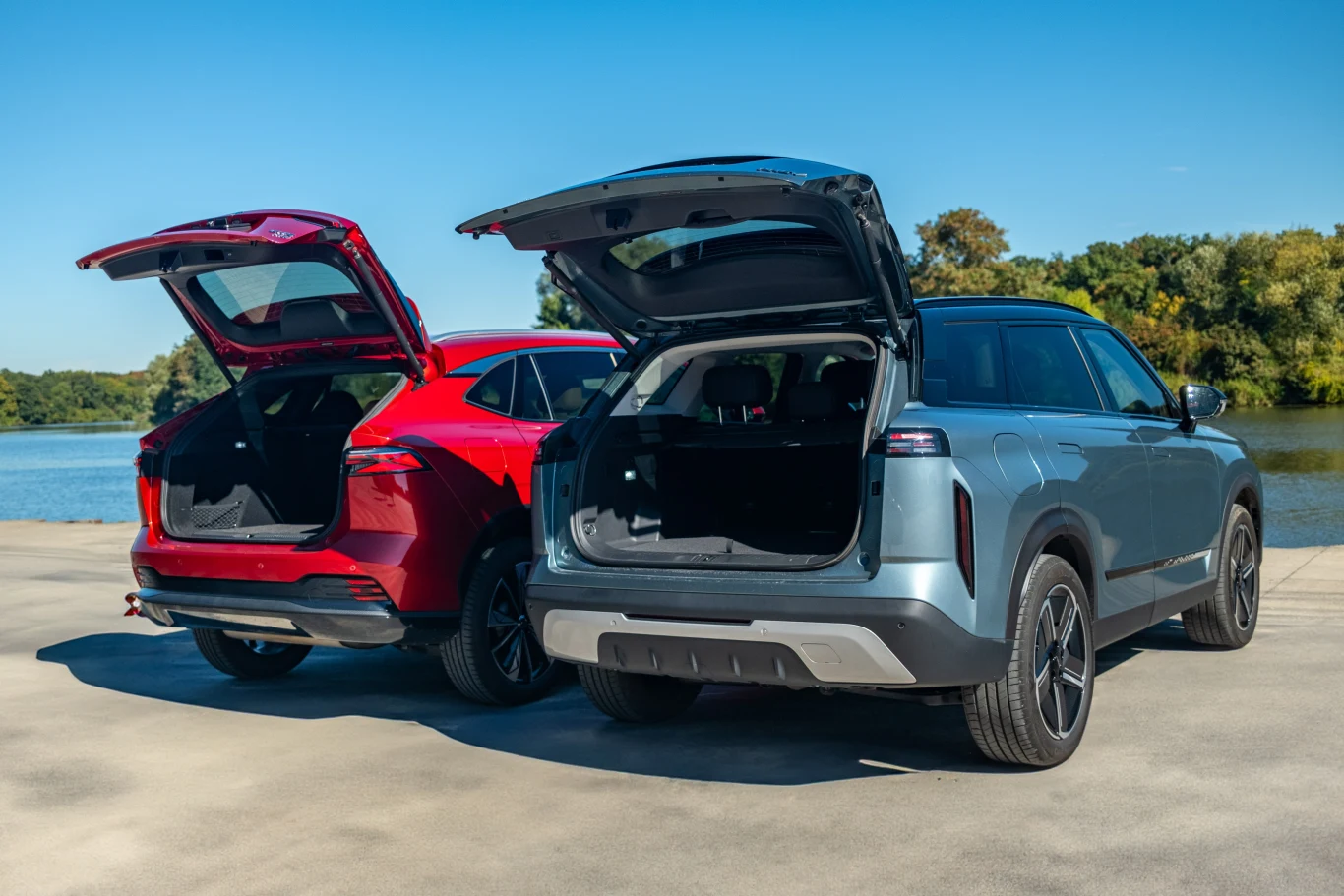 Dwa nowoczesne samochody typu SUV zaparkowane tyłem do aparatu, oba z otwartymi bagażnikami, stoją na betonowej nawierzchni nad jeziorem, w tle zieleń drzew i błękitne niebo.