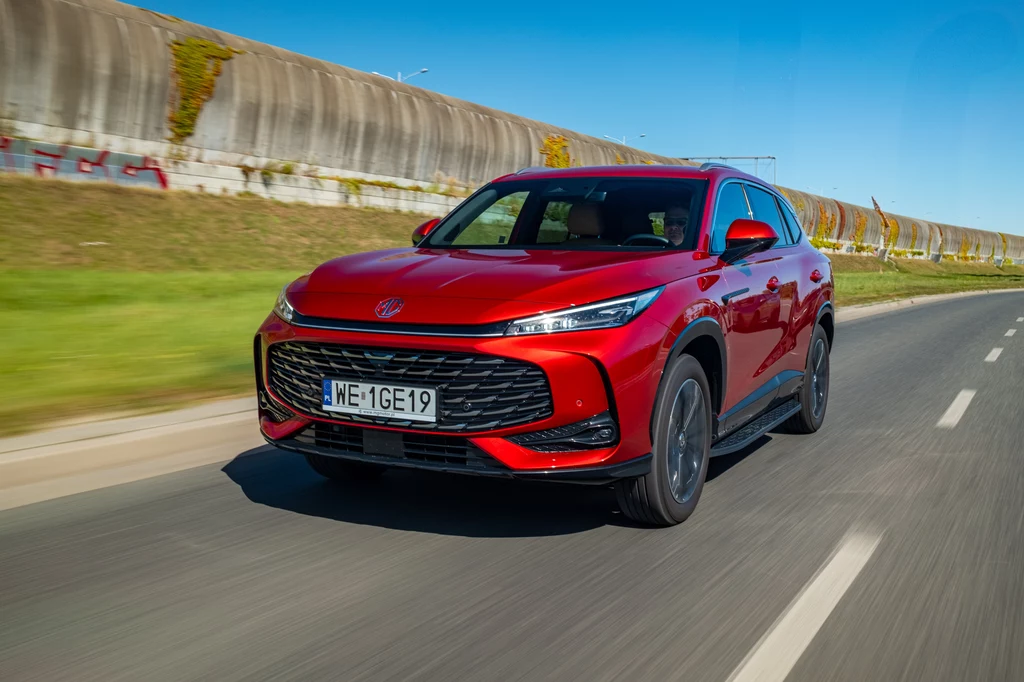 Czerwony SUV BYD jedzie po asfaltowej drodze w słoneczny dzień, mijając betonowe ekrany dźwiękochłonne oraz rząd trawy po lewej stronie.